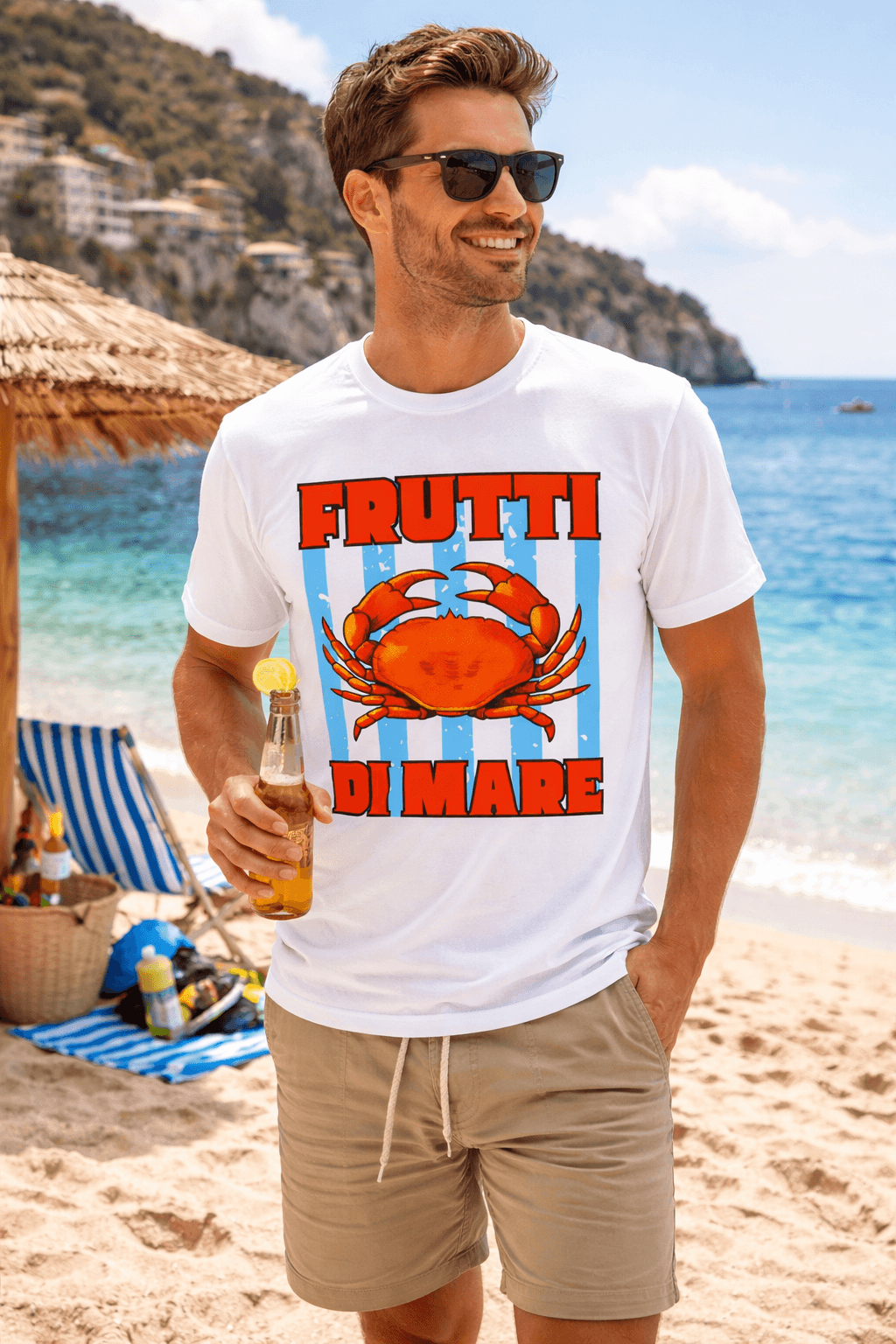 Ein junger, lachender Mann mit Sonnenbrille steht an einem sonnigen Sandstrand und hält eine Flasche mit einem Zitronenstück in der Hand. Er trägt ein weißes T-Shirt mit dem Aufdruck einer roten Krabbe auf blau-weißen Streifen und dem Text "FRUTTI DI MARE". Im Hintergrund sieht man das blaue Meer, einen Sonnenschirm und Strandliegen.